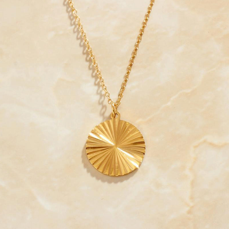 Gold Circle Sunburst Pendant Necklace sitting on a peach coloured marbled background bathed in sunlight.