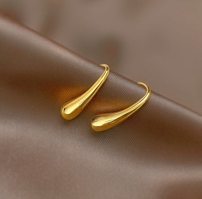 Gold Curved Droplet Earrings hanging on a dark fabric background.