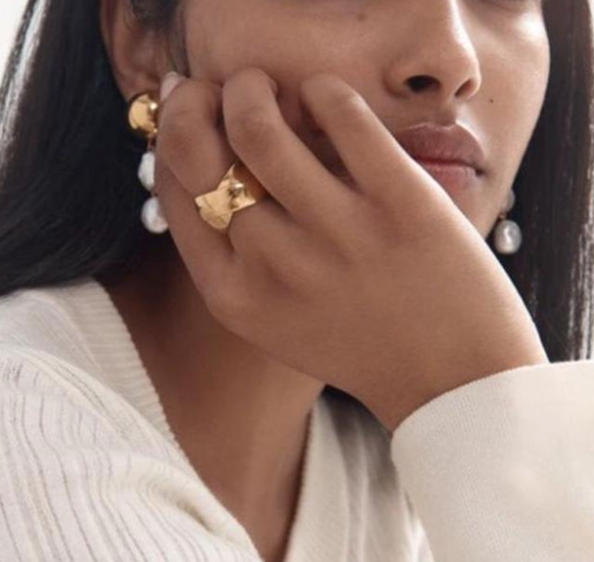 A woman in a white sweater and gold earrings, wearing interlinked gold ring looking elegant and stylish.