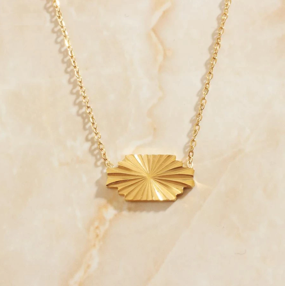 A Gold Horizontal Sunburst Pendant Necklace presented on a marbled background with warm sunlight shining on it.