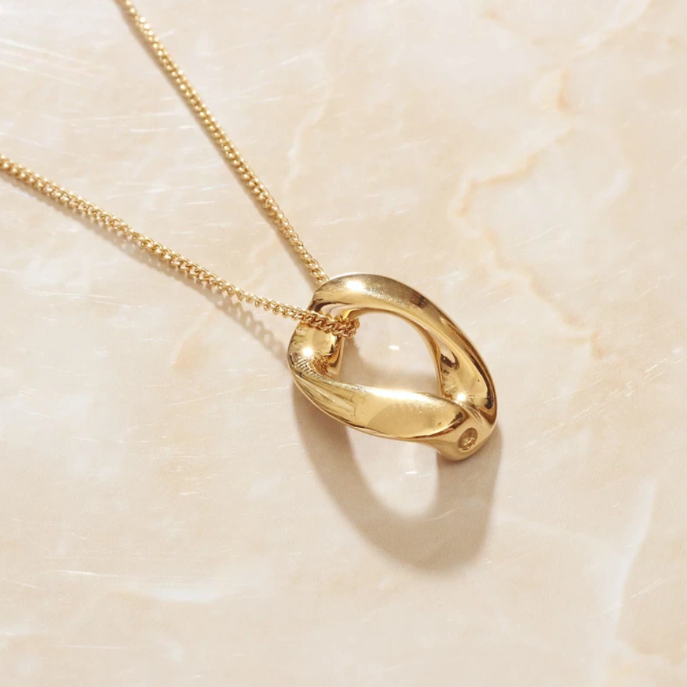 A Gold Infinite Loop Pendant Necklace presented on an angle on top of a marbled peach background