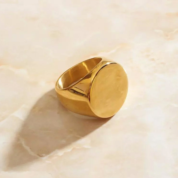 A large round faced gold signet ring is displayed on a sleek marble surface.