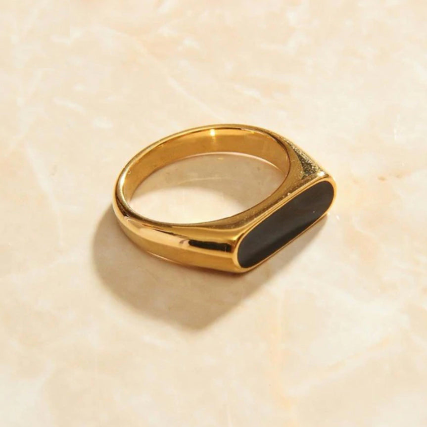 A Gold Long Black Set Signet Ring sitting on a smooth marble surface in warm sunlight.