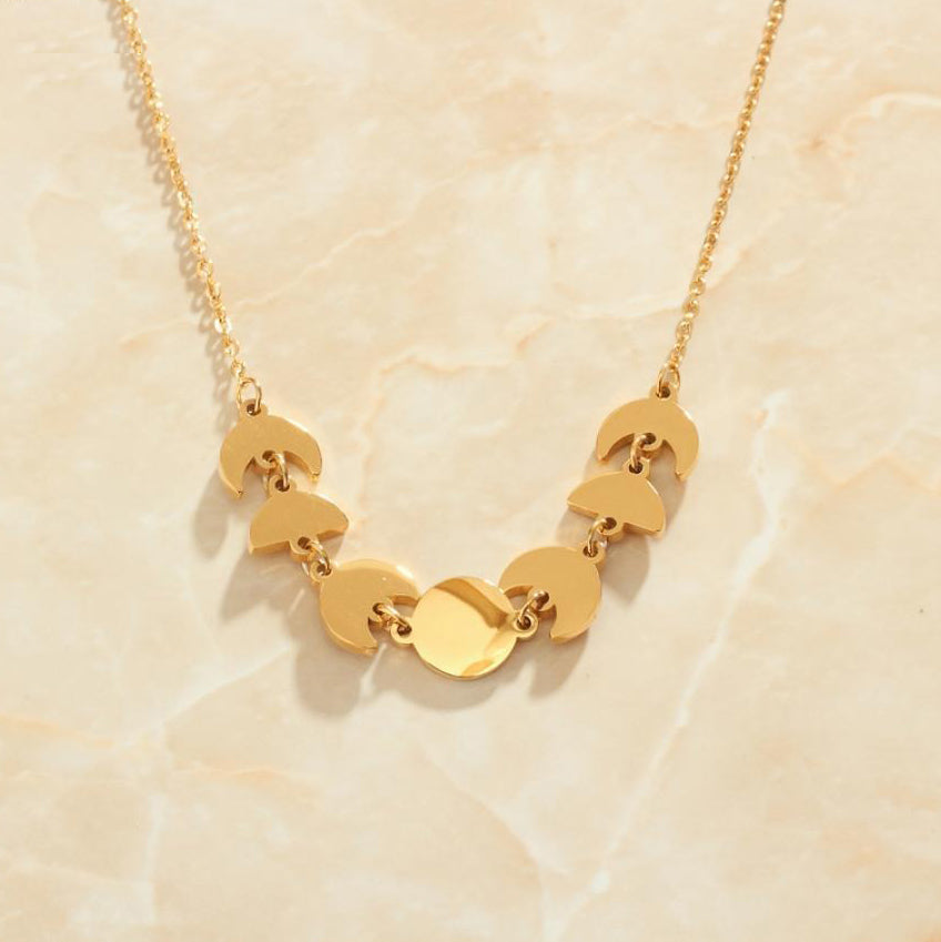 A Gold Moon Phase Necklace with the moon phase pendants hanging suspending the middle of the chain on a peach coloured marble background.