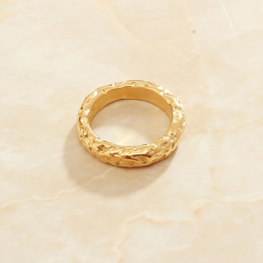 A gold ring with stone pattern laying on a sleek marble pattern which is peach in colour.