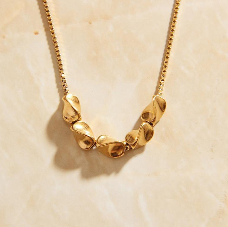 A gold boxchain necklace with five pistachio shaped pendant charms handing on the chain over a marbled background.