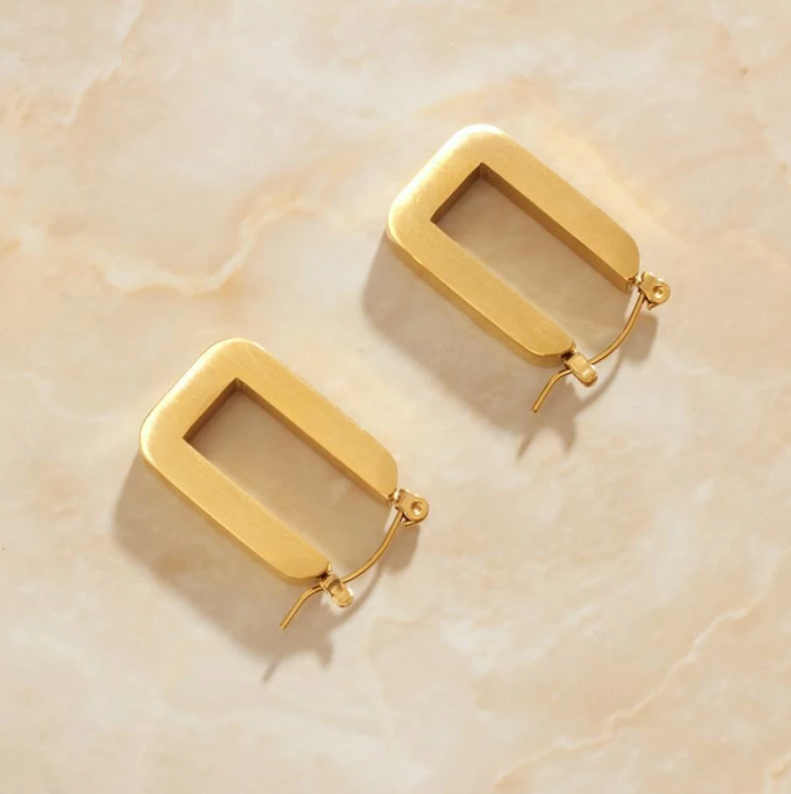 Gold Rectangular You Earrings laying on a smooth rose quartz surface shining brightly.
