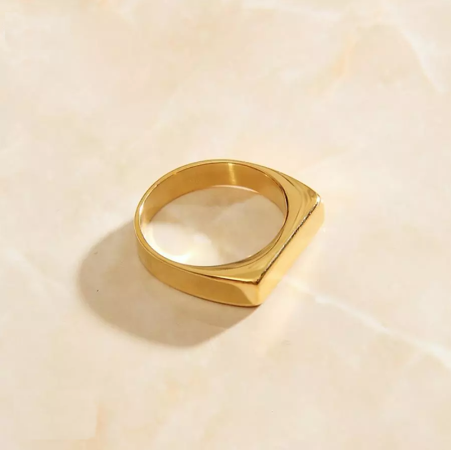 A Gold slim rectangular signet ring laying on a marble surface, showing minimalist beauty.