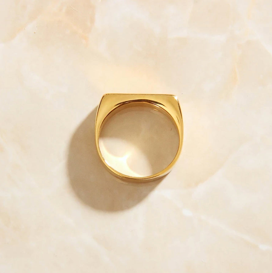 A gold slim signet ring laying on a marbled surface, shining brightly.