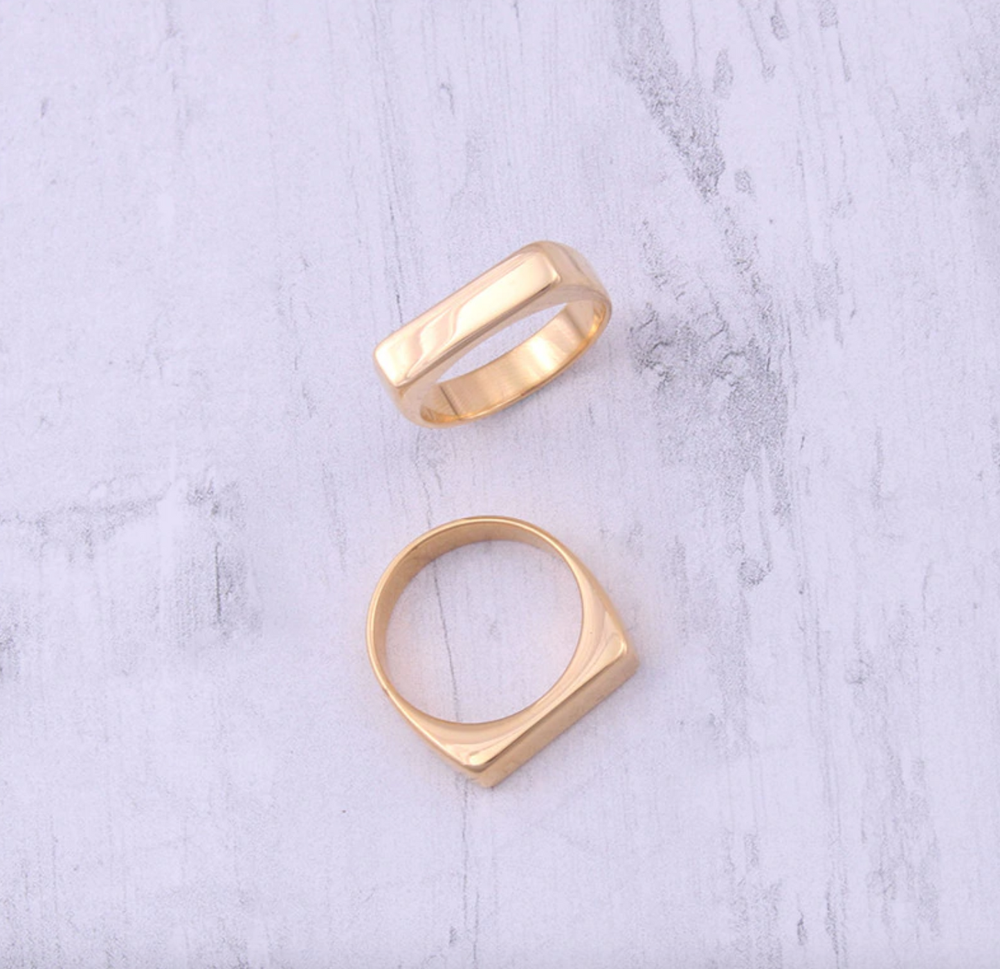 Two gold slim rectangular signet rings on a textured surface.