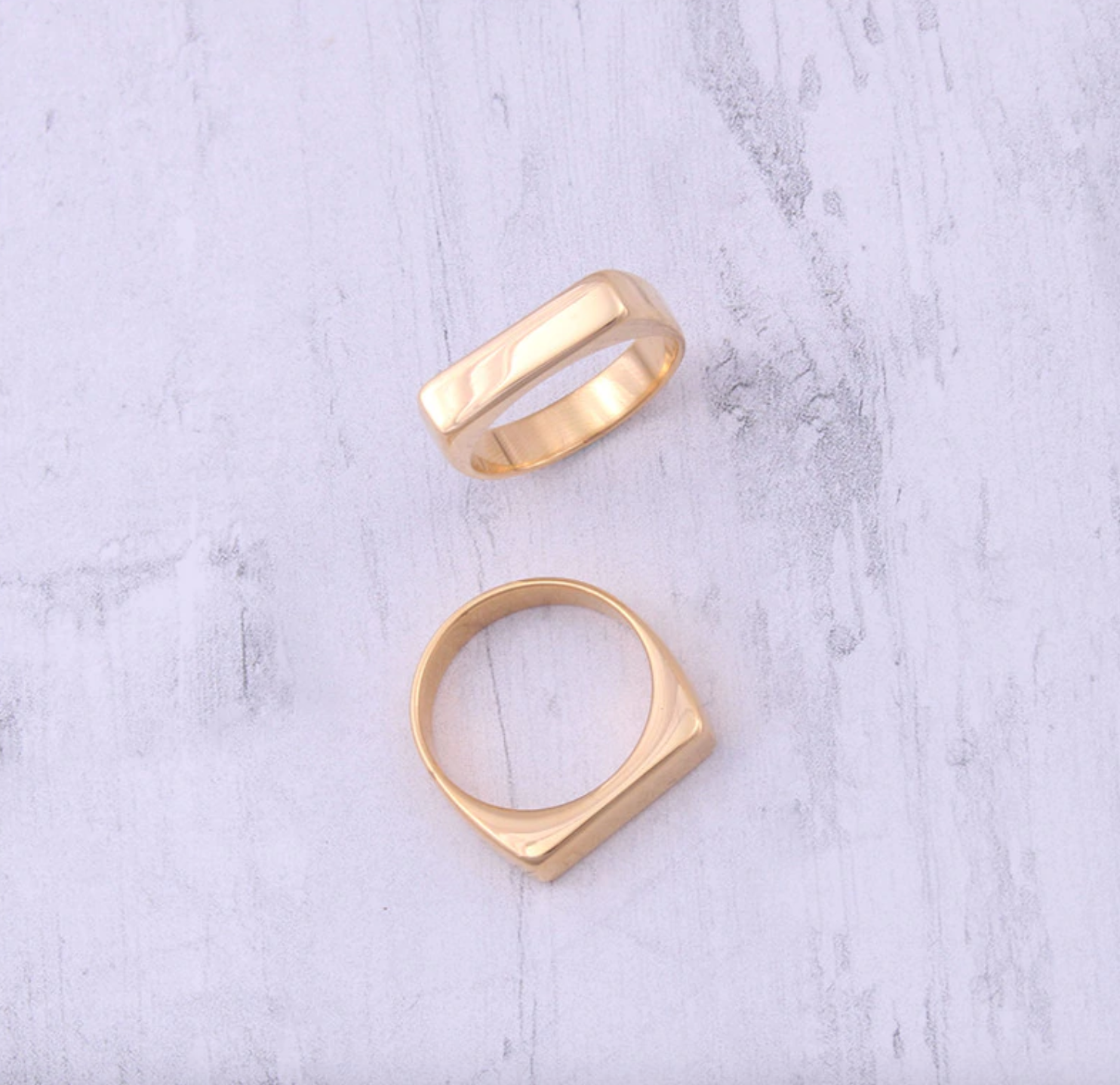 Two gold slim rectangular signet rings on a textured surface.