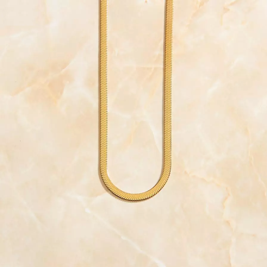 A classic minimalist Gold Snake Chain Necklace laid neatly on a marbled peach coloured background.