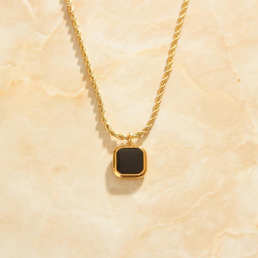 A Gold Square Black Set Pendant on a rope necklace laying on a marbled background.