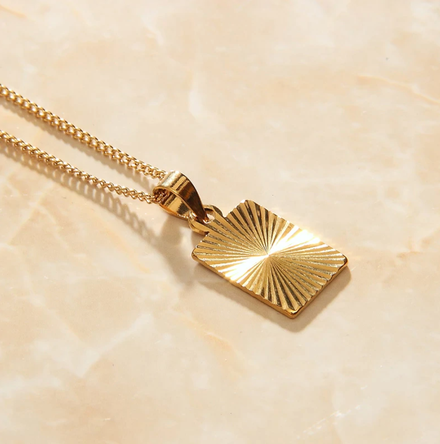 A Gold Sunburst Pendant Necklace on apricot coloured background.
