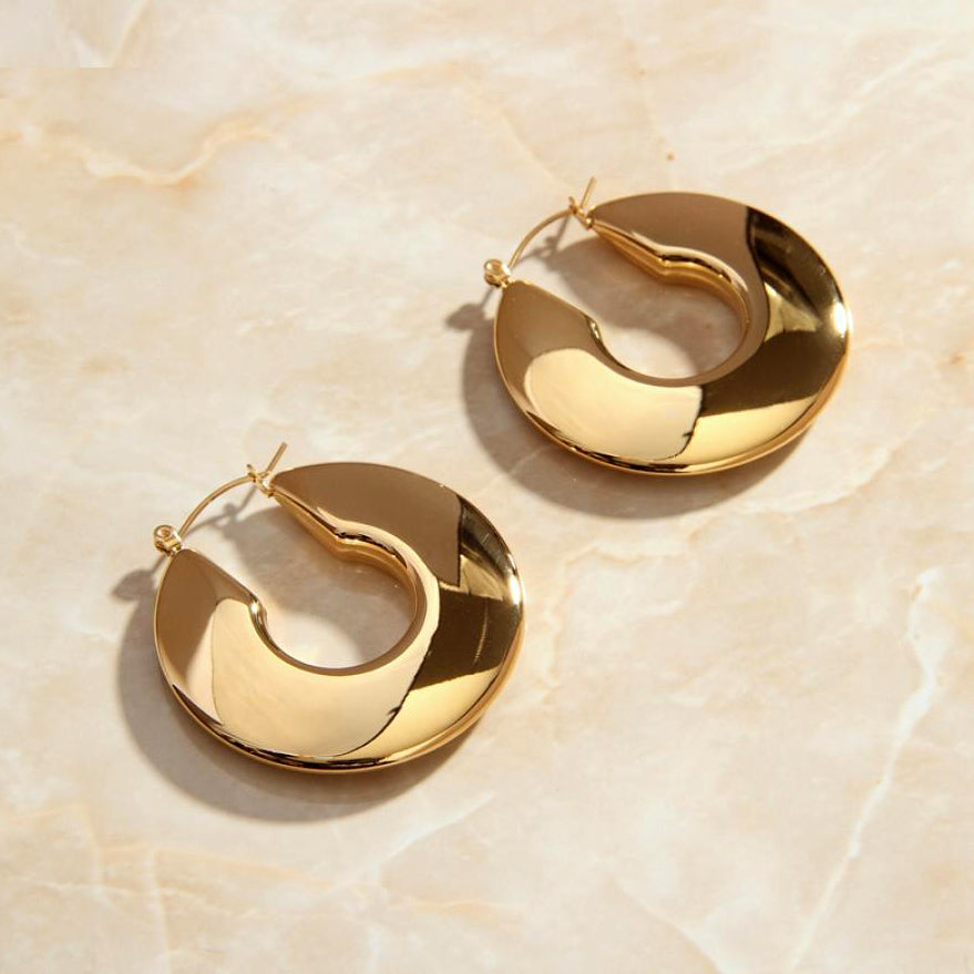 Gold Wide Polished Hoop Earrings laying on a smooth rose quartz surface shining brightly.